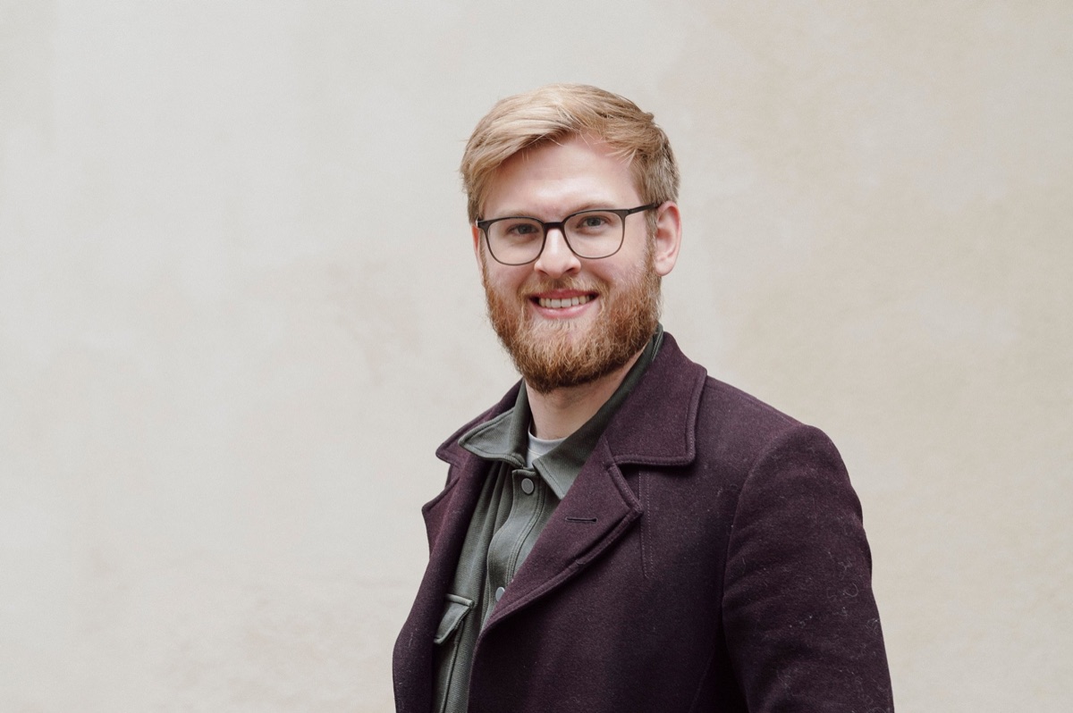 Oliver Scase, smiling, in a plum wool coat against a pale stone wall